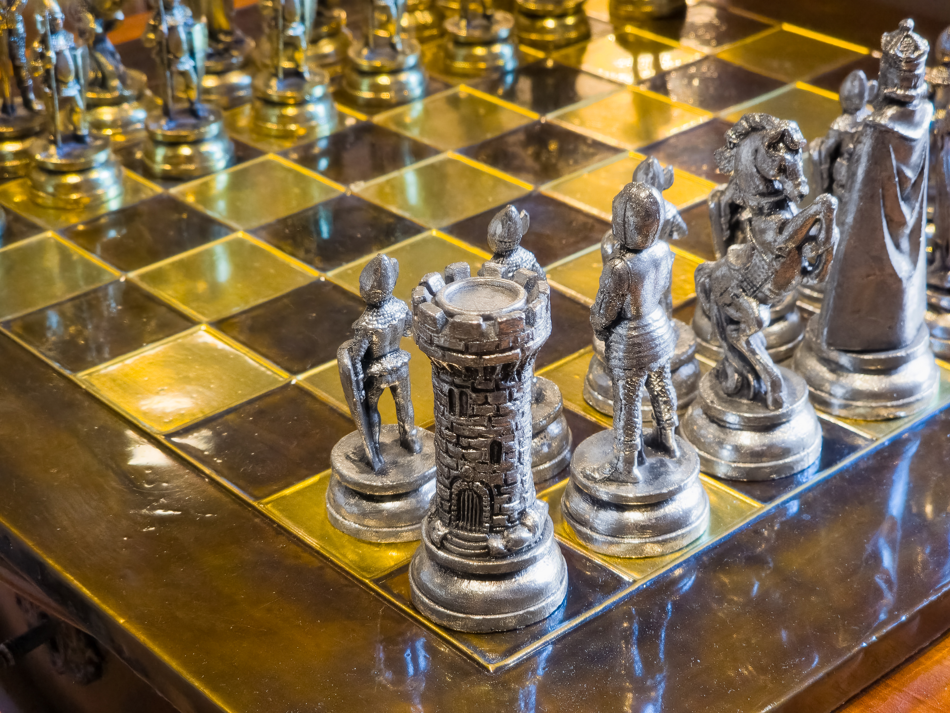 High-angle close-up of an elegant antique metal chess set with intricately detailed silver knight and rook pieces positioned on a shiny brass and bronze chessboard.